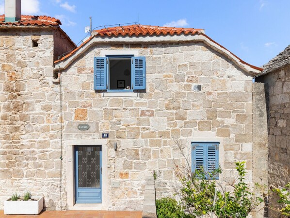 Un edificio de piedra con techo de tejas, una puerta con contraventana azul y un ventanal pequeño con contraventana azul.
