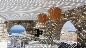 An outdoor seating area with white striped tables and chairs, stone walls, and a view of the sea. - Villa Orion Mykonos - Blue Views Mykonos Villas (Mykonos)