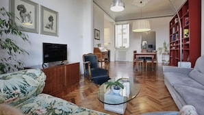 Ein Wohnzimmer mit einer Couch, einem Sessel, einem Couchtisch, einem Fernseher, einem Bücherregal und einem Essbereich mit Tisch und Stühlen.