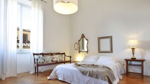 Ein Schlafzimmer mit Holzboden, einem Bett mit weißer Bettwäsche, einem Nachttisch mit Lampe, einem Spiegel an der Wand und einem Fenster mit Vorhängen.