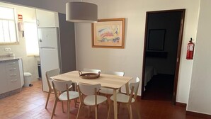 Un comedor con una mesa de madera y sillas blancas, una lámpara colgante, una cocina con armarios blancos y un extintor en la pared.
