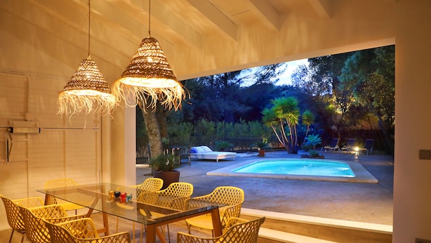 Área de refeições ao ar livre com cadeiras de vime e mesa de vidro, com vista para uma piscina e jardim.