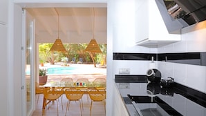Uma cozinha moderna com bancada preta e armários brancos, uma área de refeições com cadeiras amarelas e mesa de vidro, e vista para uma piscina e áreas verdes do lado de fora.
