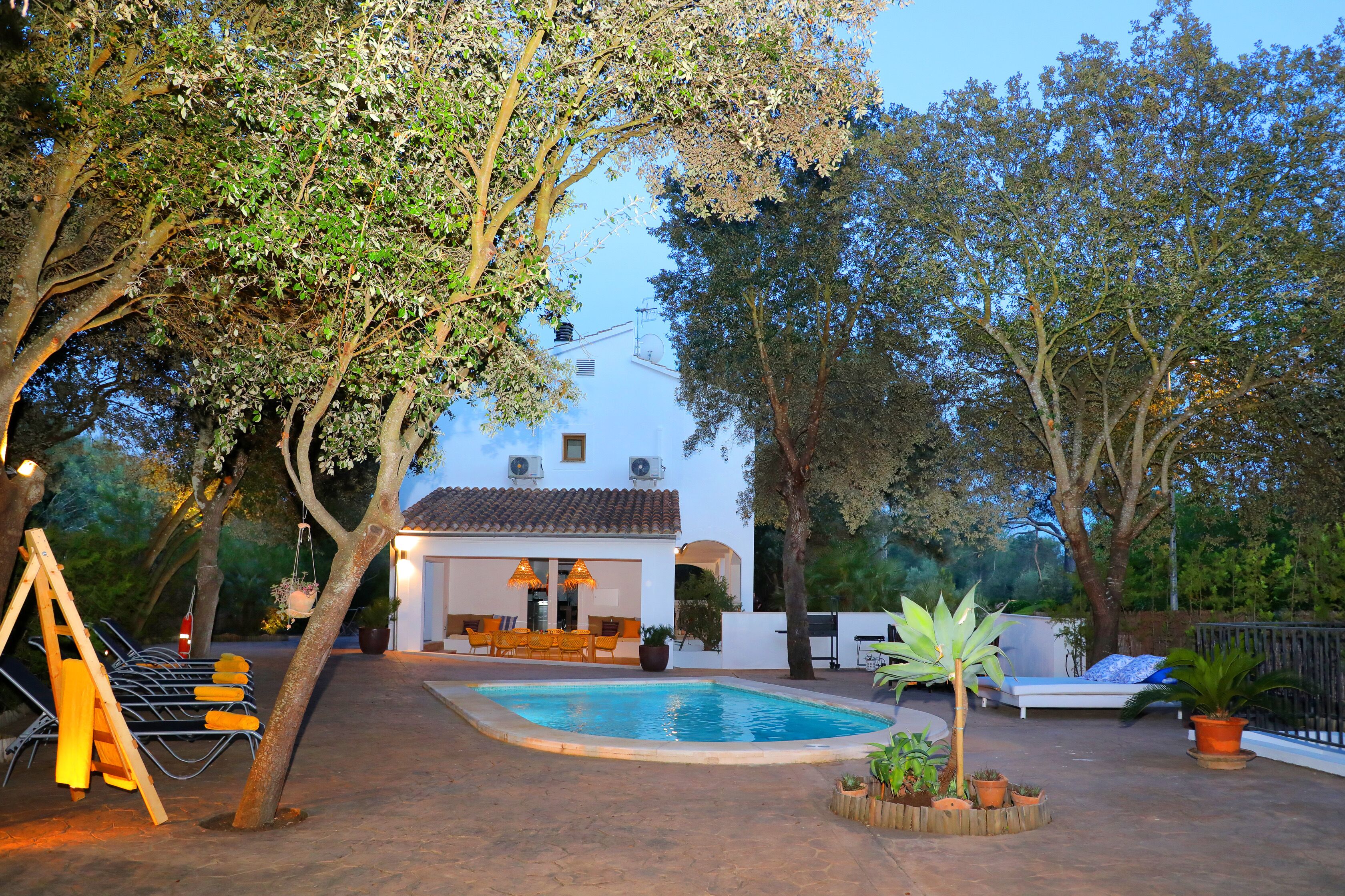 A pool area with a lounge chair, a small table, and a hanging chair. There is a white building with a tiled roof in the background.