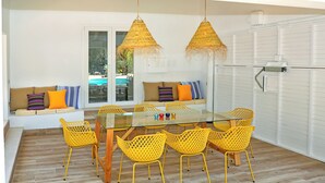 Uma área de refeições com mesa de vidro, cadeiras amarelas e um banco com almofadas coloridas.