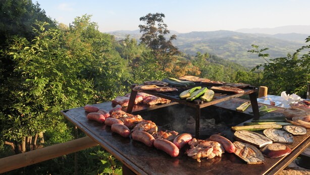 Ein Grill mit verschiedenen Fleisch- und Gemüsesorten, umgeben von üppiger Vegetation und einer Berglandschaft im Hintergrund.
