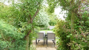 A garden patio with a table and chairs, surrounded by dense foliage and a wooden fence. - Brick and flint pet friendly cottage a short walk to sandy beach and pub (Holme next-the-sea)