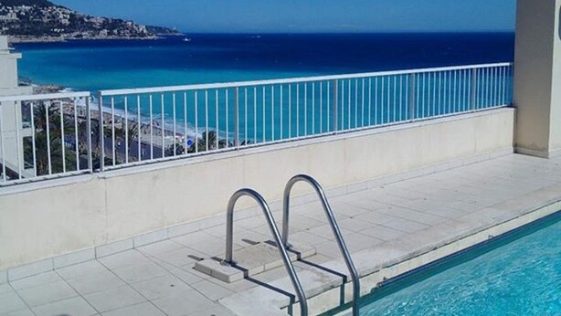 Una piscina con una escalera metálica, una barandilla blanca y vistas al océano y a una costa distante.