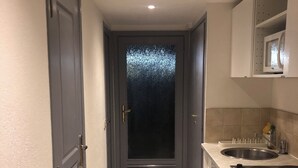 Un pasillo estrecho con una puerta oscura, una zona de cocina con fregadero y estufa, y suelo de baldosas.