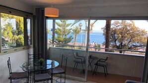 Un balcón con una mesa y sillas de vidrio, un colgante de luz y vista al mar y palmeras.