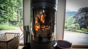 A wood-burning stove with a glass front, a fire bucket labeled "FIRE," and a woven basket on the windowsill. - Modern Cottage, big garden, AGA, Loch/Mountain Views. Cinema Room & Log Burner (Arrochar)