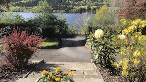 A paved pathway leading to a gate, surrounded by various plants and flowers, with a lake and forested hills in the background. - Modern Cottage, big garden, AGA, Loch/Mountain Views. Cinema Room & Log Burner (Arrochar)