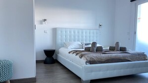 Ein modernes Schlafzimmer mit einem gebuschten Kopfteil, einem Bett mit weißer Bettwäsche und einem 'HAPPY'-Kissen, einem dunklen Nachttisch und einem hellen Hocker.