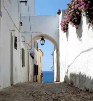 Une ruelle étroite pavée de pierres, avec un bâtiment blanc sur la droite, un grand arc et une vue sur la mer au loin.