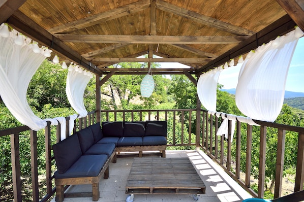 A covered outdoor seating area with a wooden bench and a sofa, surrounded by white curtains and a wooden railing.