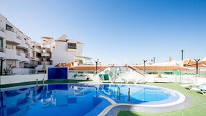 Piscina con agua azul cristalina, rodeada por un césped verde y edificios blancos con balcones.