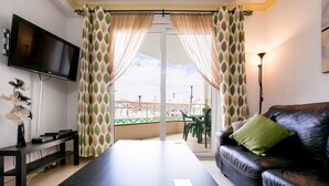 Una sala de estar con sofá de cuero negro, una mesa de centro, televisor de pantalla plana montado en la pared y un balcón con vistas a la ciudad.