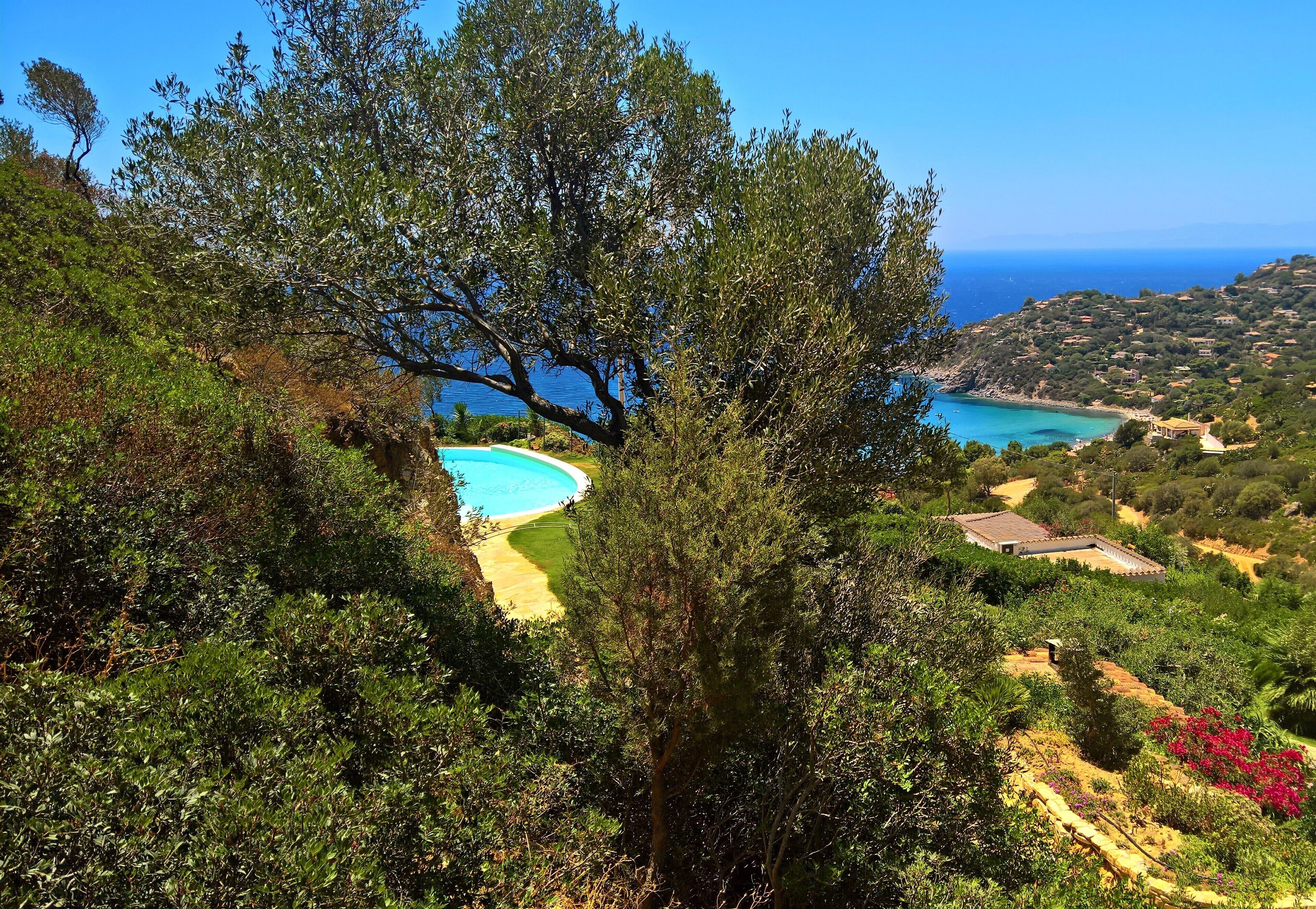 A coastal landscape with a swimming pool, lush greenery, and distant views of the sea and hills.