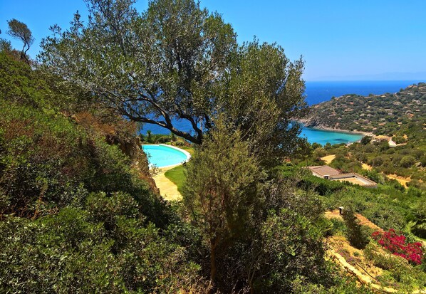 A coastal landscape with a swimming pool, lush greenery, and distant views of the sea and hills.