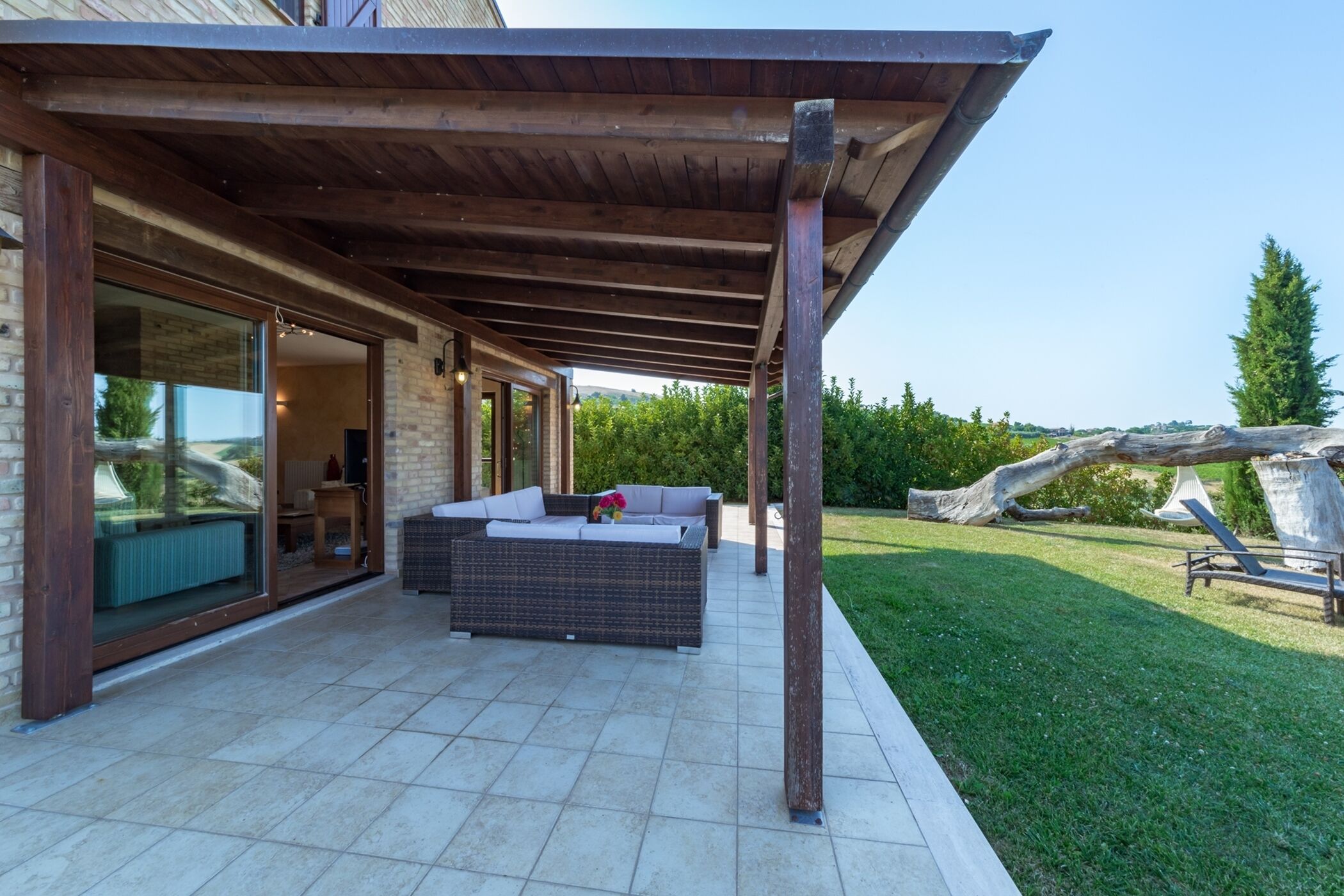 A covered patio with a wicker sofa, a glass door leading inside, and a view of a grassy area with a large tree and a sculpture.
