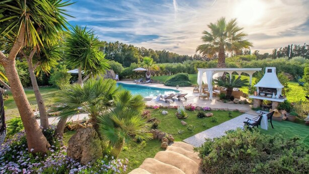Ein Poolbereich mit Liegestühlen, einer Pergola und einem Grillplatz, umgeben von Grünflächen und Palmen.