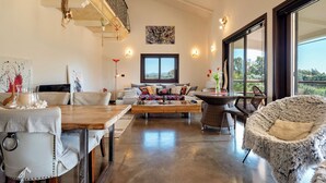 Ein geräumiges Wohnzimmer mit Couchtisch aus Holz, einer Couch mit bunten Kissen, einem Esstisch mit Stühlen und einem großen Fenster mit Blick ins Freie.