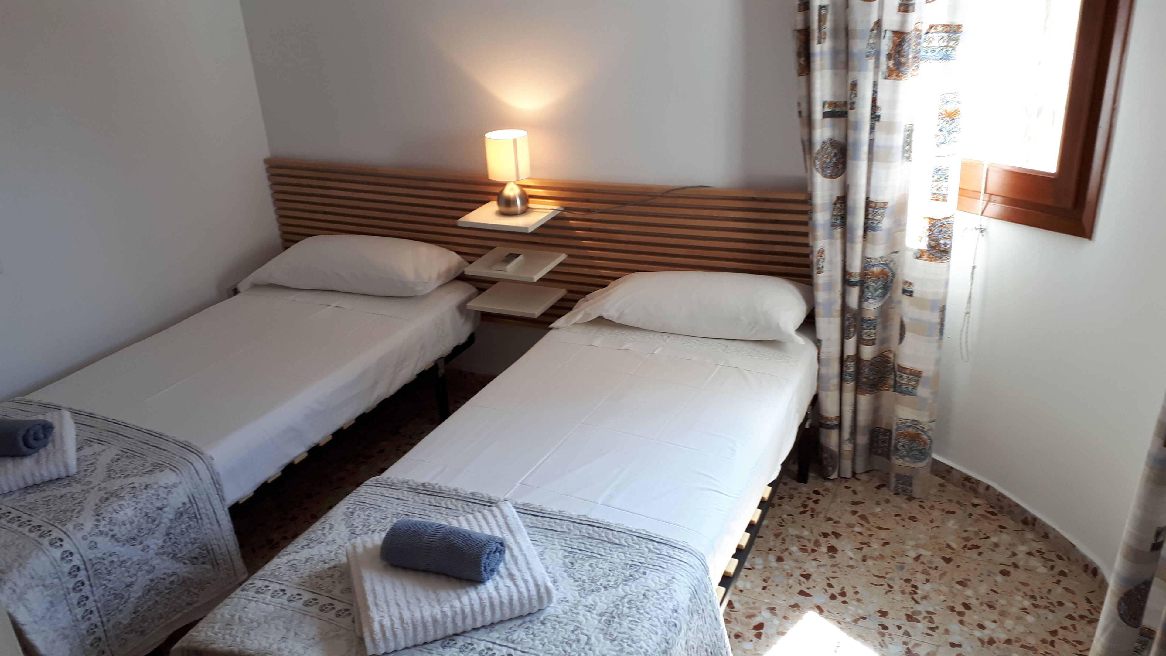 Habitación de hotel con dos camas individuales, una mesita de noche con lámpara, una alfombra estampada y una ventana con cortinas.