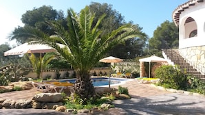 Zona de piscina con sillones de descanso y sombrillas, rodeada de vegetación y un edificio de piedra.
