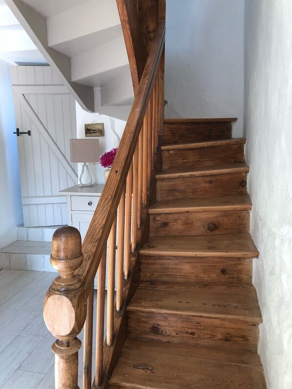 Una escalera de madera con barandilla que lleva a un rellano con una puerta blanca, una mesita con una lámpara y flores.