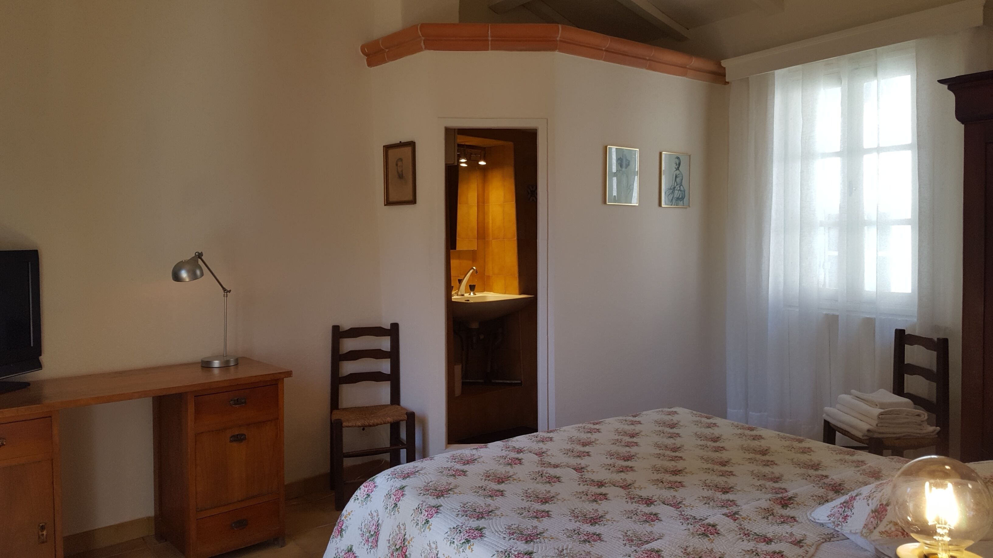 Quarto com escrivaninha de madeira, cadeira, cama com colcha florida, mesa de cabeceira com abajur e janela com cortinas.