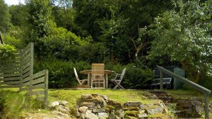A rustic outdoor seating area with a wooden table and chairs, surrounded by a stone wall and lush greenery.