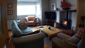 A cozy living room with a sofa, armchair, and a wooden coffee table. A television is mounted on the wall, and a fireplace is visible in the background.