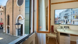 Een kamer met een groot raam met uitzicht op een historisch gebouw, een bureau met stoel en een lijst met een afbeelding aan de muur.