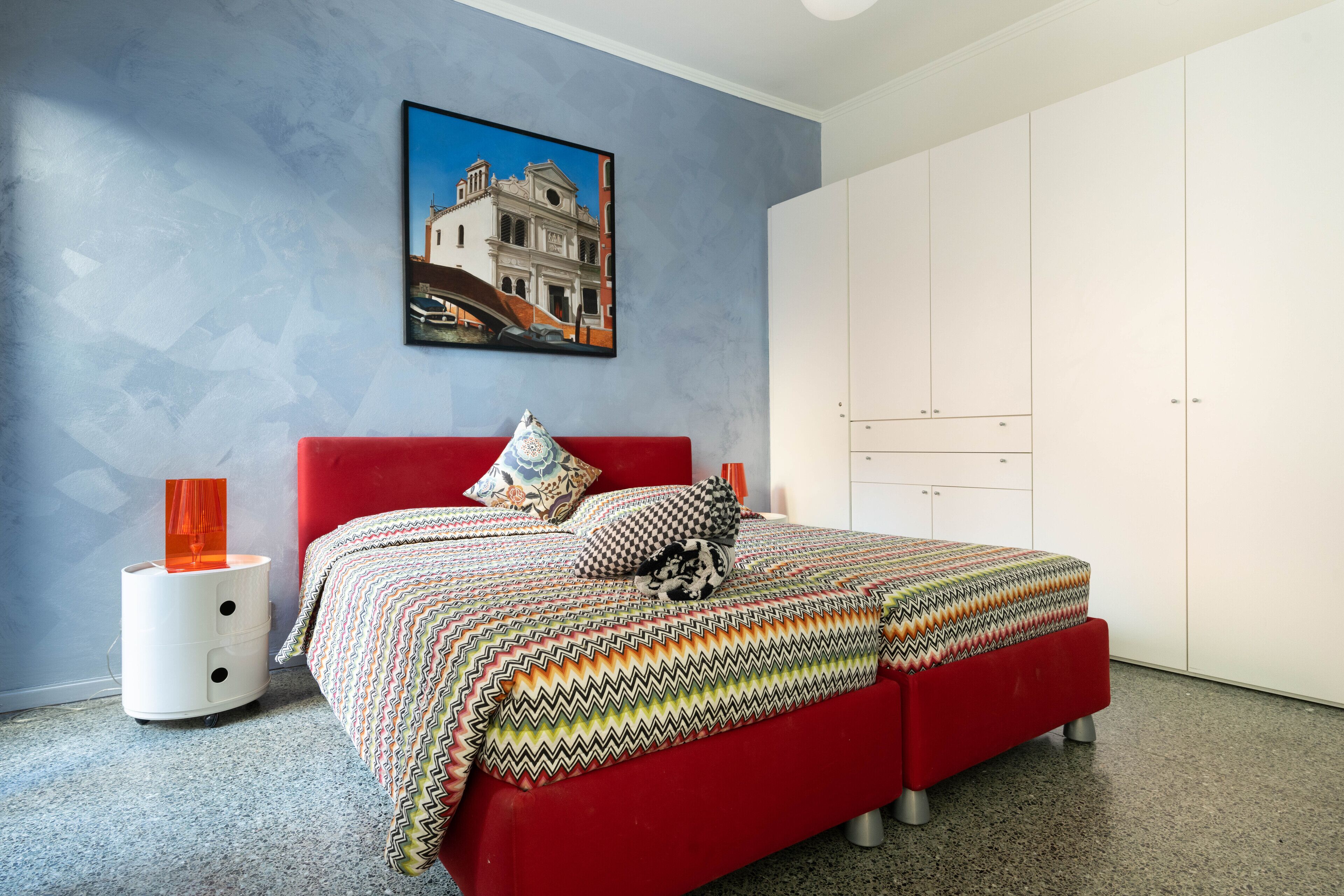 Une chambre avec un lit rouge, une armoire blanche et un tableau accroché au mur.