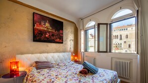 Een slaapkamer met een groot bed, een schilderij van een stadsgezicht, uitzicht op gebouwen door het raam en een radiator.