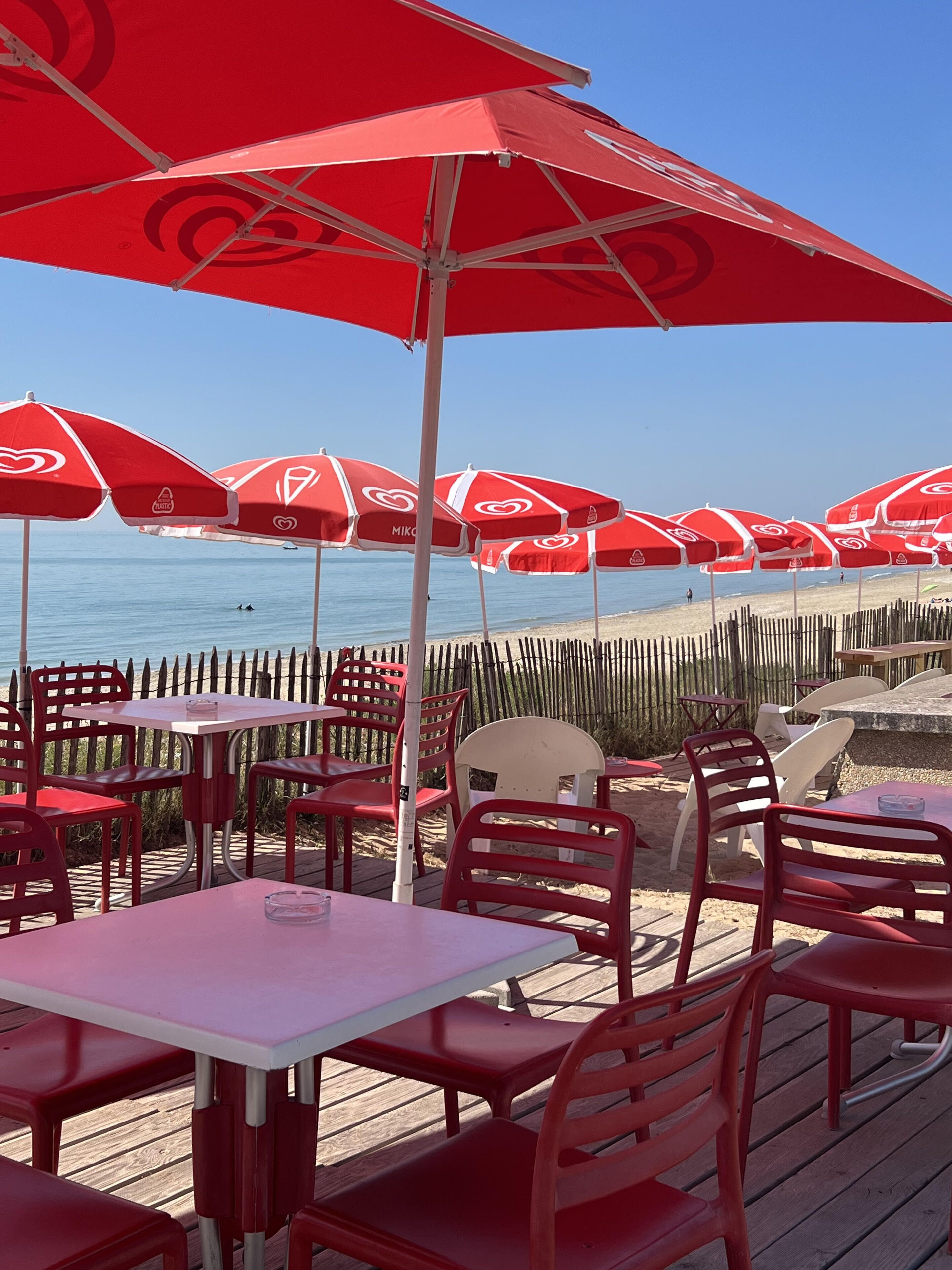 Espace extérieur aménagé avec des parasols rouges, des tables blanches et des chaises rouges, offrant une vue sur la plage.