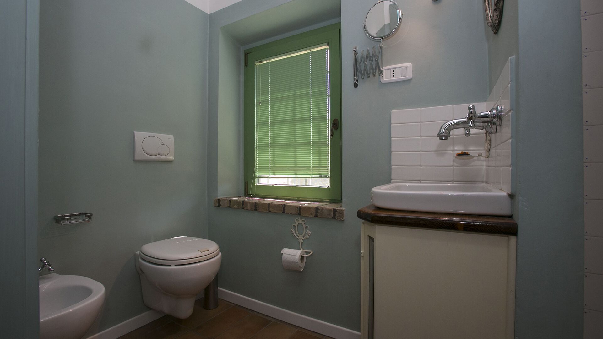 A bathroom with a toilet, bidet, sink, and a window with blinds.