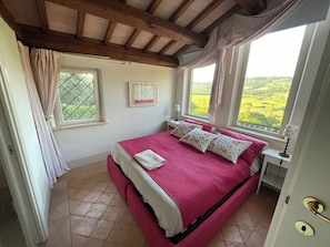 A bedroom with a bed, a window, a nightstand, and a view of the countryside.