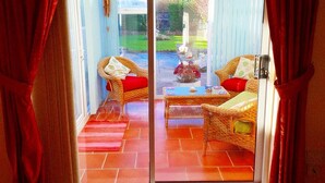 A room with a glass door leading to an outdoor area. Inside, there are wicker chairs with red cushions and a small table with a vase and a bowl.
