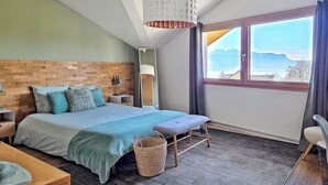 Une chambre moderne avec une tête de lit en bois, un grand lit recouvert d’une couverture verte, une table de chevet avec une lampe, une vue sur les montagnes par la fenêtre et un petit banc.