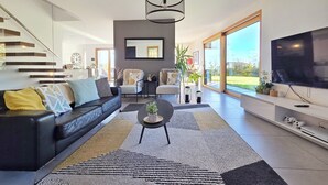 Un salon moderne avec un canapé en cuir noir, une table basse et un grand téléviseur. Un escalier se trouve sur la gauche, et une porte-fenêtre coulissante donne accès à un jardin.