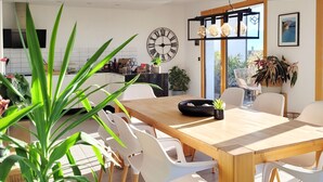 Un espace repas moderne avec une table en bois et des chaises blanches, agrémenté de plantes en pot et d’une grande fenêtre donnant sur l’extérieur.