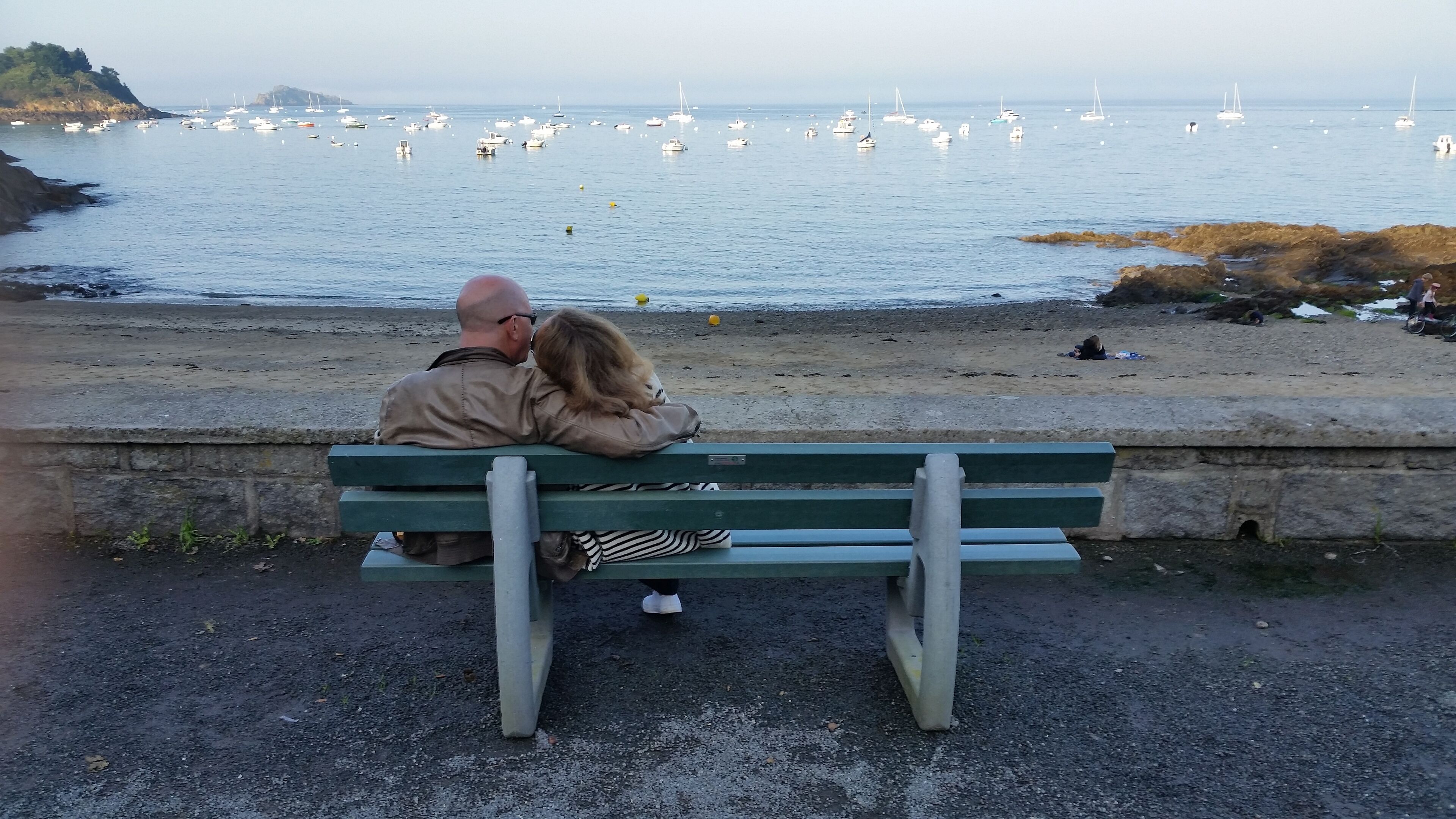 Ein Paar sitzt auf einer Bank und blickt auf ein ruhiges Meer mit Booten und einer felsigen Küste.