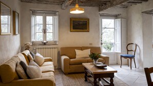 A cozy living room with a sofa, a coffee table, and a chair. There are windows with curtains, a painting on the wall, and a hanging light fixture. - Torretta - Luxury rustico with pool and air conditioning EV wallbox (Camaiore)