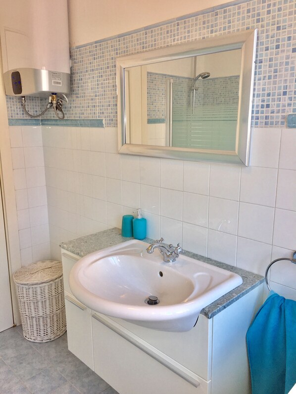 A bathroom with a white sink, a mirror, and a shower. There is a towel hanging on the wall and a basket on the floor. - Trieste Panoramic gulf view (Trieste)