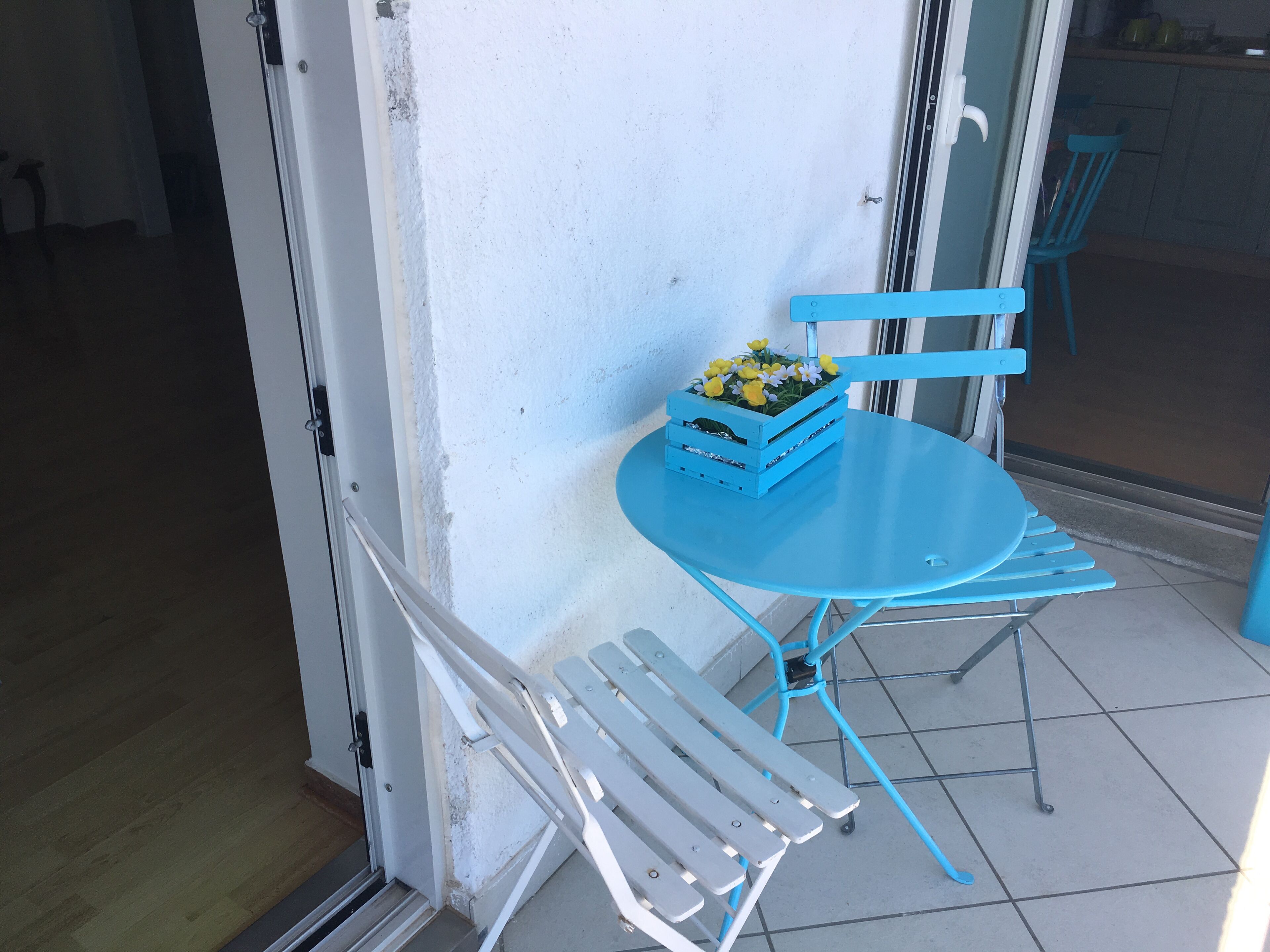 A blue metal table and chairs with a blue crate containing flowers on a tiled patio.