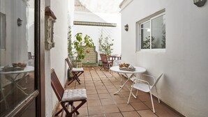 Une cour étroite avec un mur blanc, un sol carrelé, une petite table ronde entourée de chaises et une fenêtre.