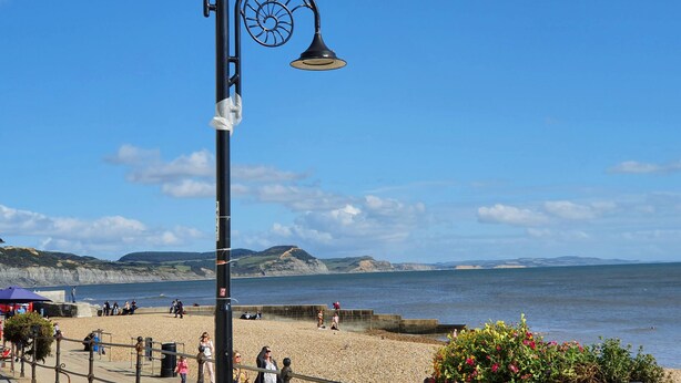 A coastal promenade with a pebbled beach, a promenade railing, and a clear view of the sea and distant cliffs.