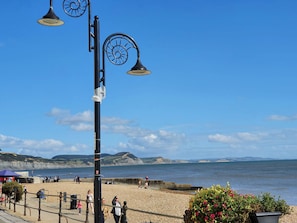 A coastal promenade with a pebbled beach, a promenade railing, and a clear view of the sea and distant cliffs.