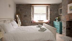 A bedroom with a stone wall, a bed with white bedding, a wooden window seat, and a teal dresser.